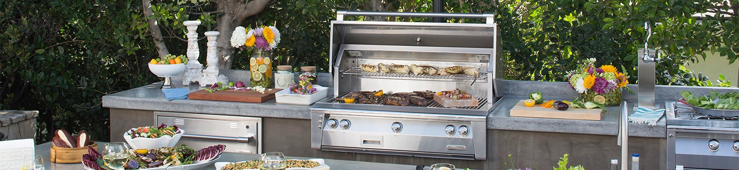 Outdoor kitchen with a stainless steel grill filled with various grilled foods, surrounded by fresh vegetables, dishes, flowers, and a sink, creating a vibrant and functional outdoor cooking space.