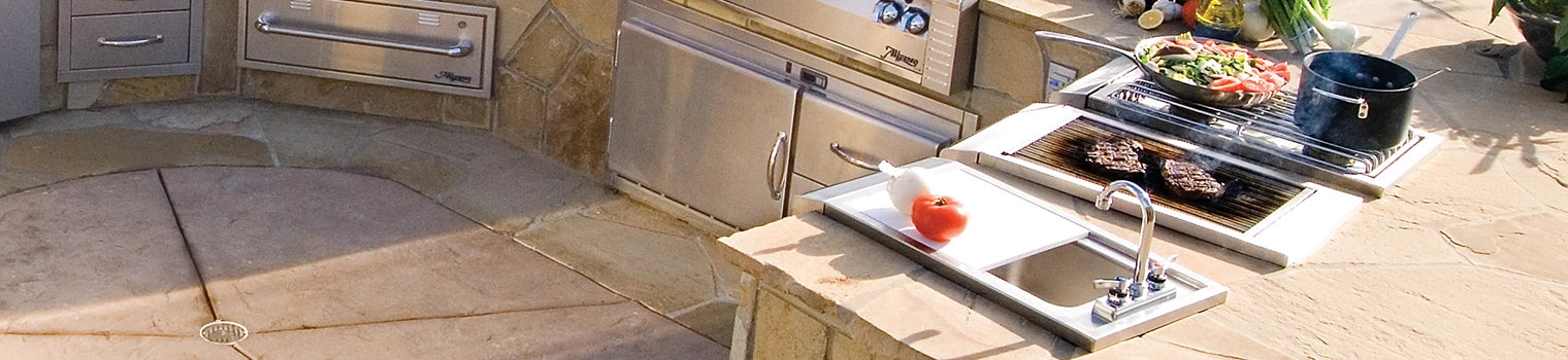 Outdoor kitchen setup with a built-in grill cooking steaks, a sink, and food preparation areas featuring grilling accessories and ingredients