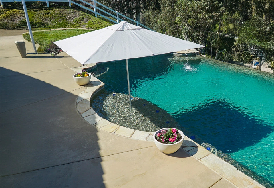 Large white commercial patio umbrella providing shade by a luxurious poolside, featuring a sparkling infinity pool and vibrant flower planters for a resort-style ambiance