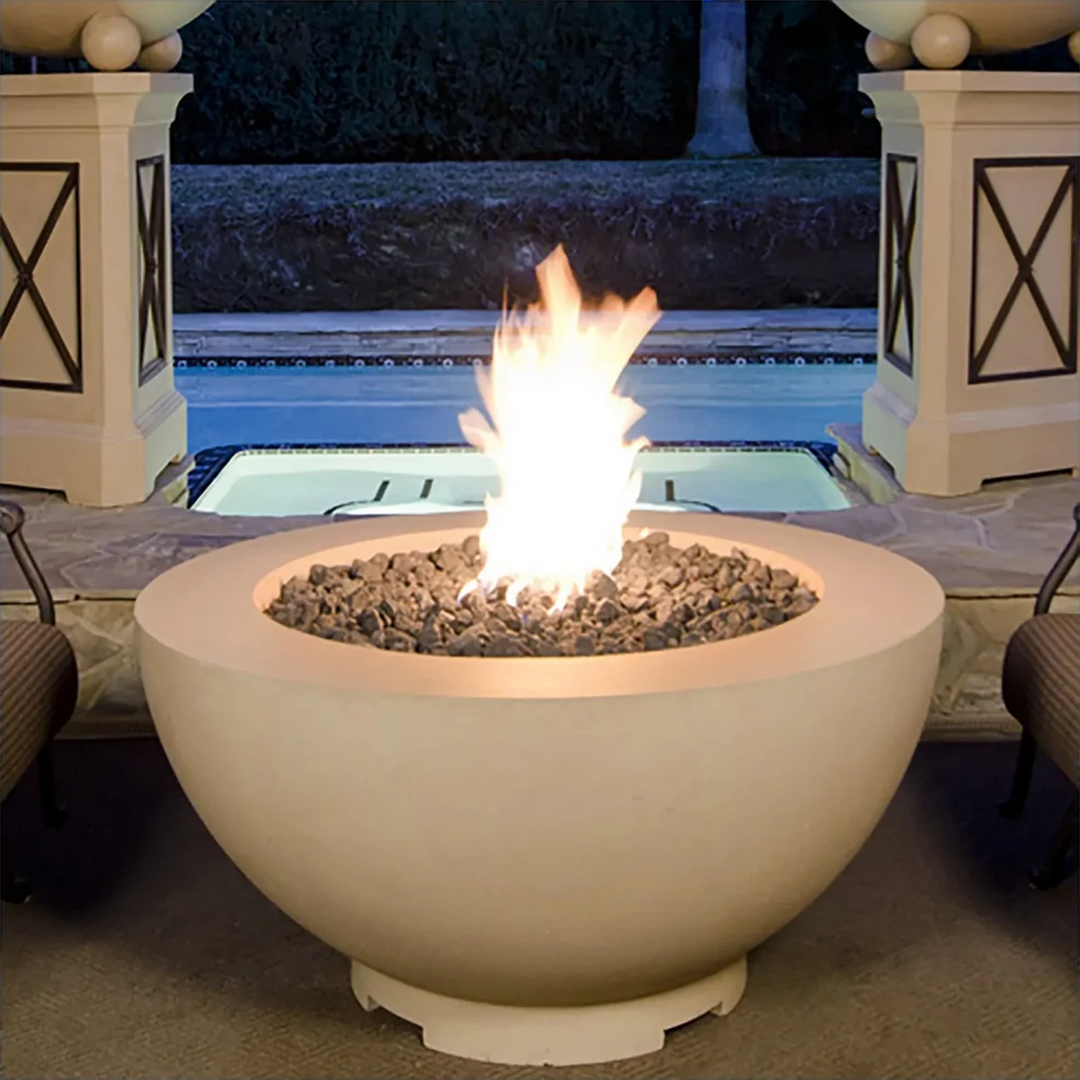 Large round beige outdoor fire pit with burning flame and lava rocks, situated on a patio area near a swimming pool with decorative columns in the background, during nighttime