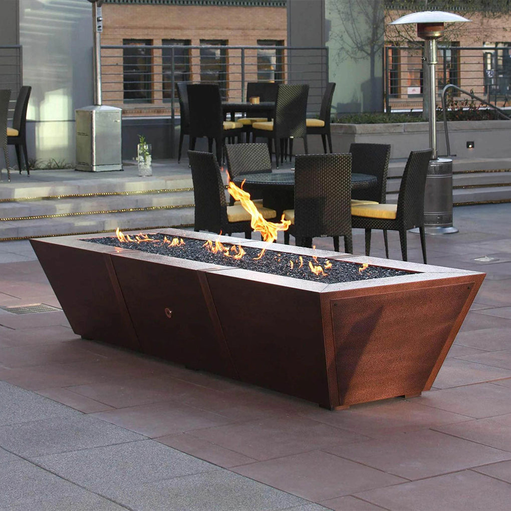 Modern outdoor fire pit with a sleek design in a commercial patio setting, featuring a long rectangular shape and flames burning on top of fire glass, surrounded by dining tables and chairs.