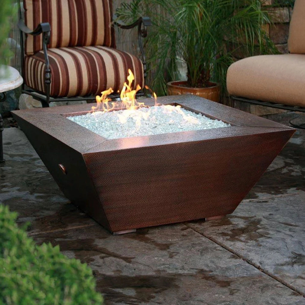 Square copper finish outdoor fire pit with flames burning over clear fire glass, placed in a cozy patio setting with cushioned outdoor furniture and greenery