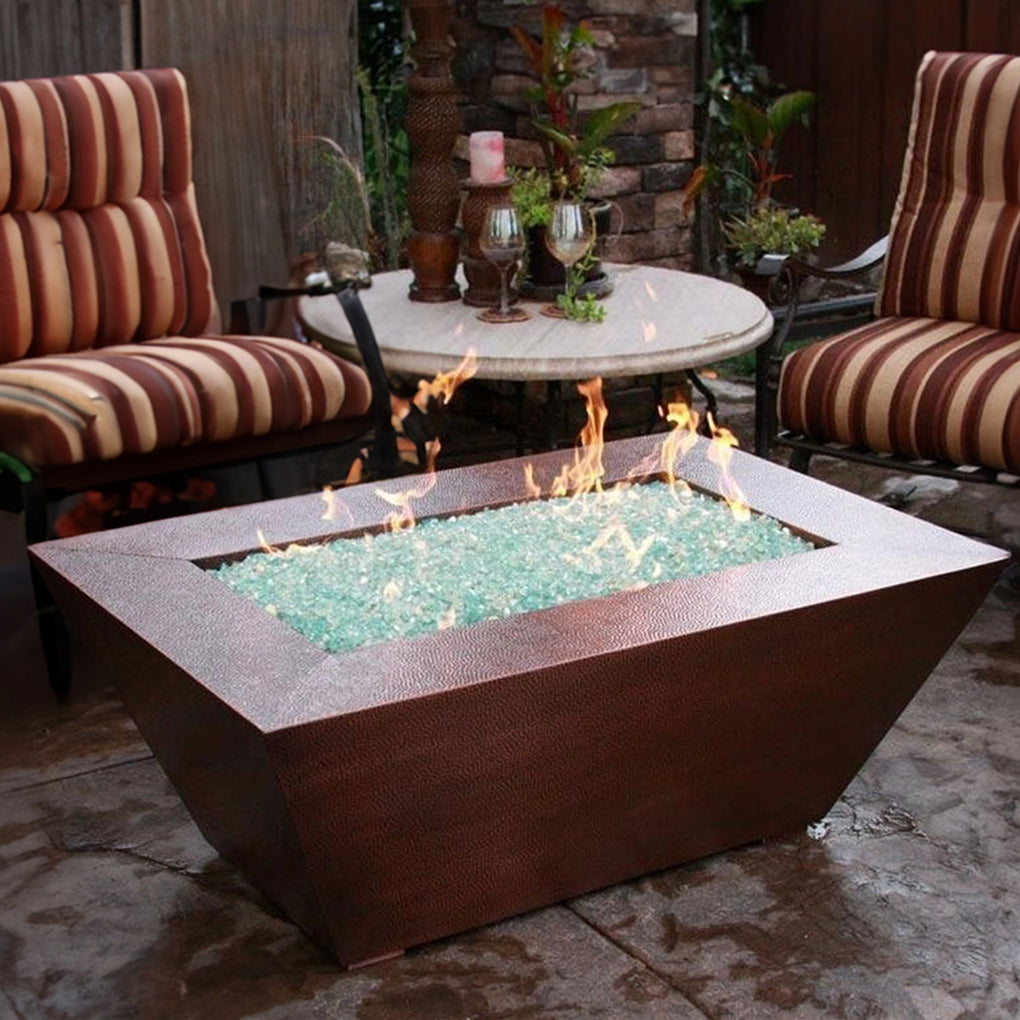 Rectangular copper finish outdoor fire pit with light blue fire glass and flames, surrounded by striped cushioned armchairs and a round table with candles in a cozy patio setting with greenery