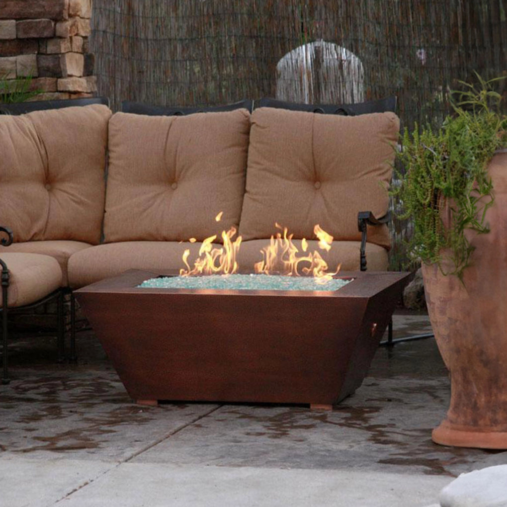 Square copper fire pit with a textured surface and small flames, placed on a patio with a beige tufted sectional sofa and a rustic stone and bamboo backdrop