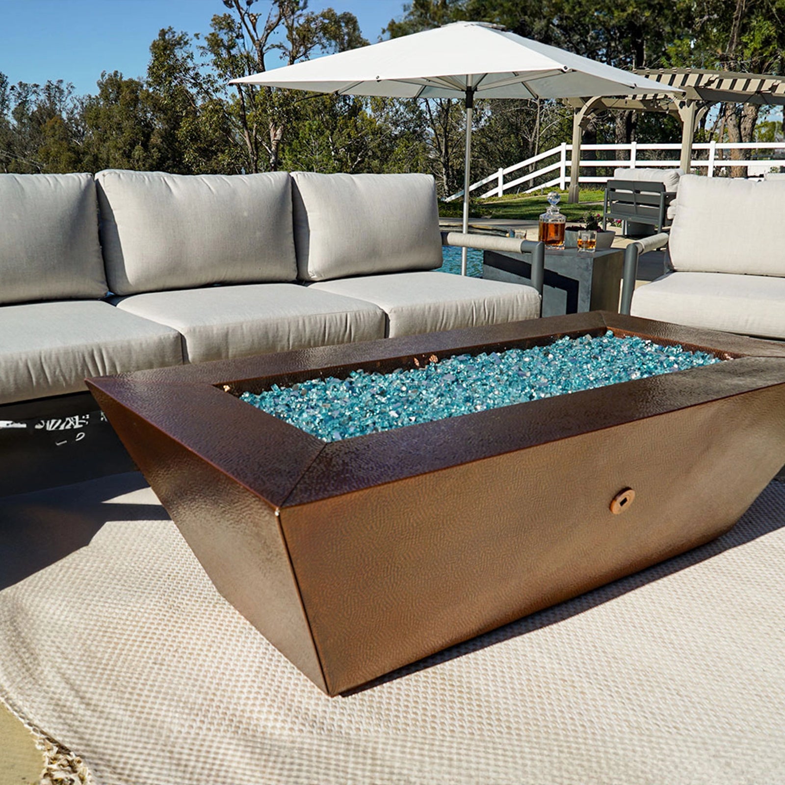 Rectangular copper finish outdoor fire pit with light blue fire glass on a beige rug, surrounded by light grey cushioned seating, an umbrella, and a scenic view of trees and a white fence