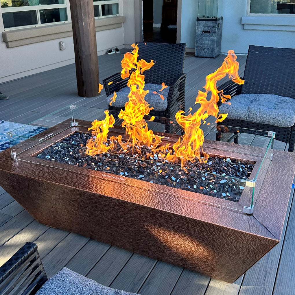 Rectangular copper fire pit with a textured surface, tall flames, and dark fire glass, set on a wooden deck with outdoor wicker chairs in the background