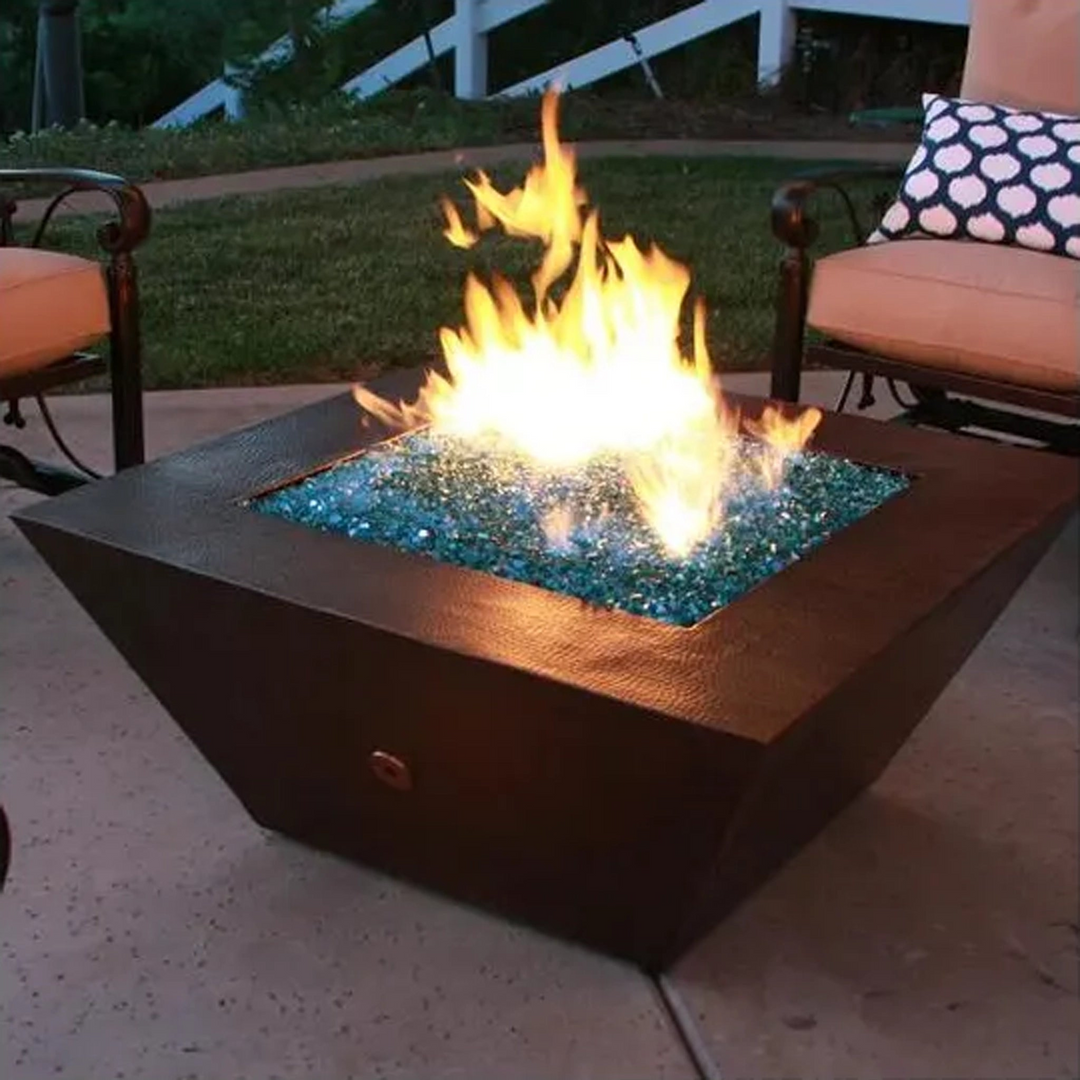 Modern square fire pit filled with blue fire glass, burning brightly on a patio with outdoor seating in the background