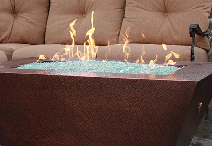 Close-up of a copper fire pit filled with turquoise fire glass, with flames rising in front of a beige cushioned outdoor sofa, creating a cozy patio ambiance