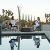 The EcoSmart Fire Ark 40 Graphite round gas fire pit enhances a desert-inspired poolside setting where two women are enjoying drinks. This sleek fire pit offers a contemporary and sophisticated touch to any outdoor area. Made from durable concrete, its design complements modern landscaping, making it a perfect fire feature for luxury poolside lounges, patios, and high-end backyard spaces.