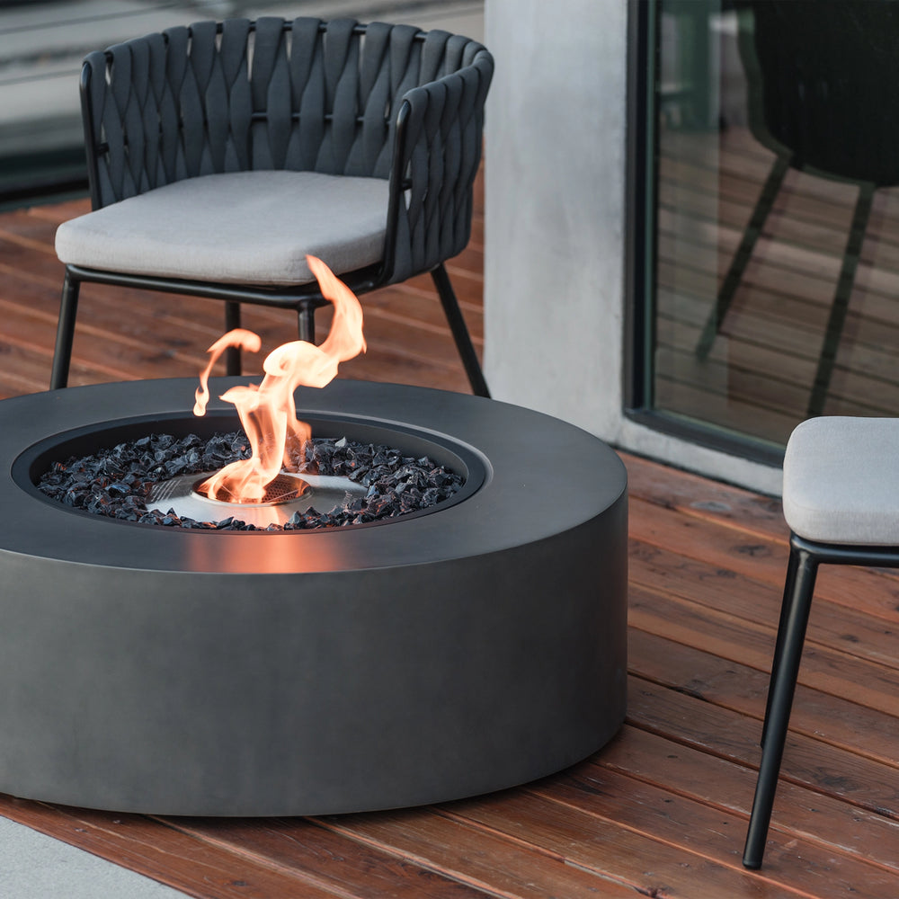 A close-up of the EcoSmart Fire Ark 40 Graphite round fire pit, elegantly placed on a wooden deck with stylish woven chairs. Featuring a stainless steel burner and black fire glass, this fire feature exudes contemporary sophistication. Ideal for patios, decks, and luxury backyard settings, this gas fire pit is perfect for creating a cozy outdoor ambiance.