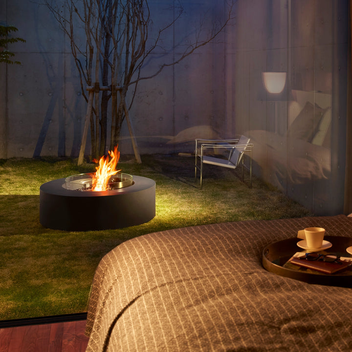 The EcoSmart Fire Ark 40 Graphite round fire pit is showcased in a modern outdoor bedroom setting. This contemporary fire feature, made of durable materials, is positioned on a grassy area outside a glass-walled bedroom, creating a warm and inviting ambiance. Ideal for luxury outdoor spaces, it features a black burner and a protective glass wind guard. Perfect for backyards, patios, and outdoor fire pit setups.