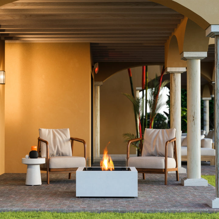 A beautifully styled EcoSmart Fire Base 30 outdoor fire pit is set on a luxurious patio, surrounded by modern wooden armchairs with neutral cushions. The fire pit table features a sleek rectangular design with a tempered glass wind guard, enhancing the ambiance of this elegant outdoor space. Its contemporary aesthetic makes it a perfect centerpiece for backyard fire pits, patio fire tables, and modern outdoor gatherings.