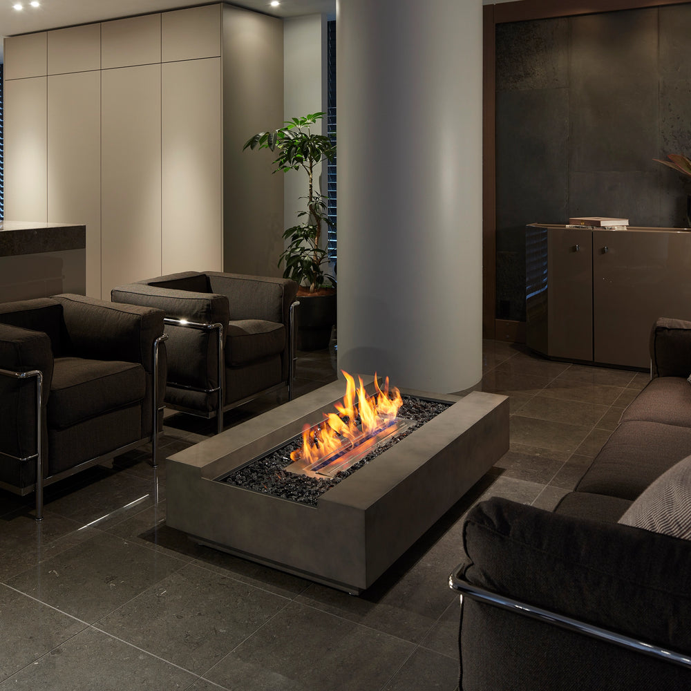 The EcoSmart Fire Cosmo 50 indoor fire table in a commercial setting, creating a cozy atmosphere in a luxury lounge. Its rectangular gas fire pit design is complemented by a sleek concrete finish, black fire media, and a wide flame display. This modern outdoor fire pit doubles as an elegant centerpiece for hospitality spaces, offering warmth and sophistication for indoor and outdoor use.