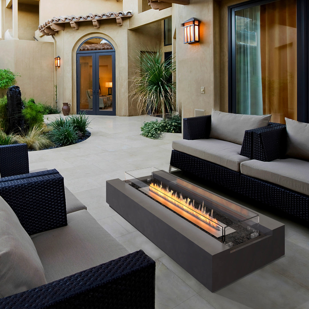 A contemporary outdoor patio featuring the EcoSmart Fire Cosmo 50 in a dark concrete finish. This luxury fire pit is surrounded by modern lounge seating, blending seamlessly into the sophisticated courtyard setting. With its rectangular gas fire pit design and tempered glass wind guard, it offers a high-end outdoor fire experience, perfect for residential and commercial applications.