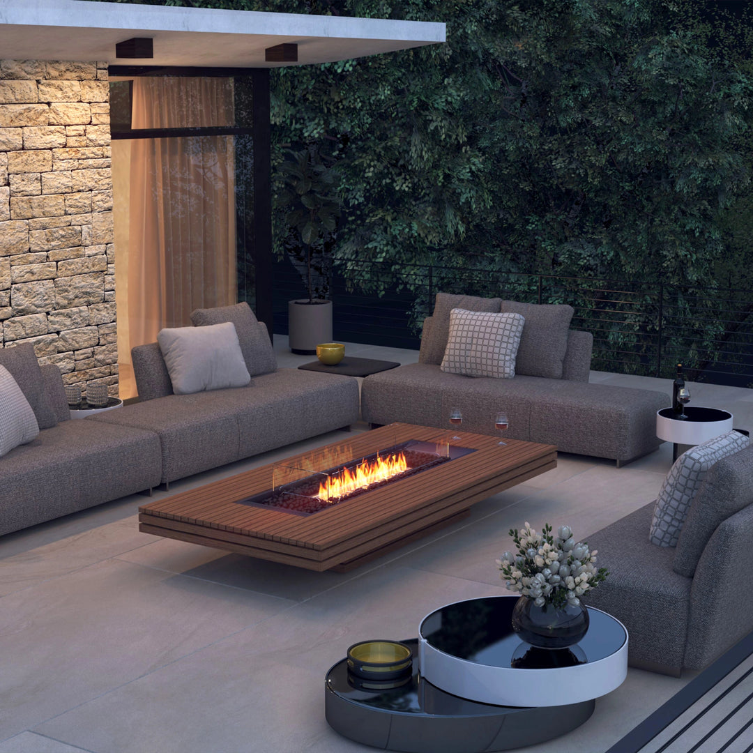 The EcoSmart Fire Gin 90 (Low) fire table in a cozy outdoor lounge area at dusk. Featuring teak wood slats and a glass wind guard, this modern fire pit provides a sophisticated focal point among plush sectionals. The warm natural gas flames add a cozy touch, ideal for backyard entertaining and outdoor relaxation.