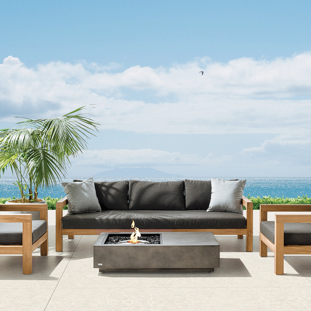 A breathtaking outdoor gas fire pit setup with the EcoSmart Fire Manhattan 50 on a spacious seaside patio. The modern fire table is paired with wooden-framed sofas and palm plants, creating a tranquil, stylish retreat. Ideal for luxury fire pits in resorts, homes, or commercial spaces, offering warmth and ambiance for entertaining guests.