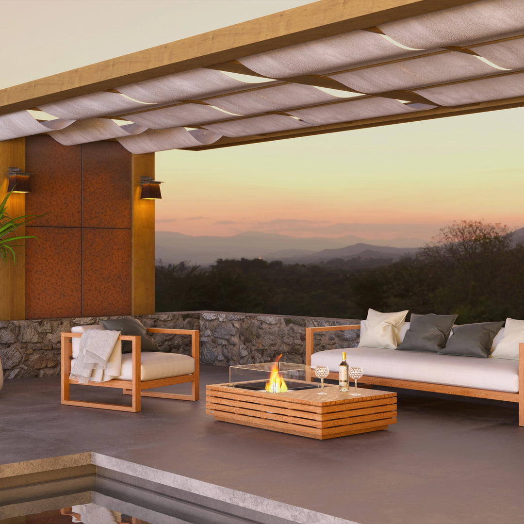 A stunning outdoor fire pit setup featuring the EcoSmart Fire Manhattan 50 teak fire table on a luxury patio overlooking a scenic sunset. Surrounded by elegant teak wood furniture, the propane fire table adds warmth and ambiance, making it a perfect fire pit for outdoors on patios, pool decks, and garden spaces.