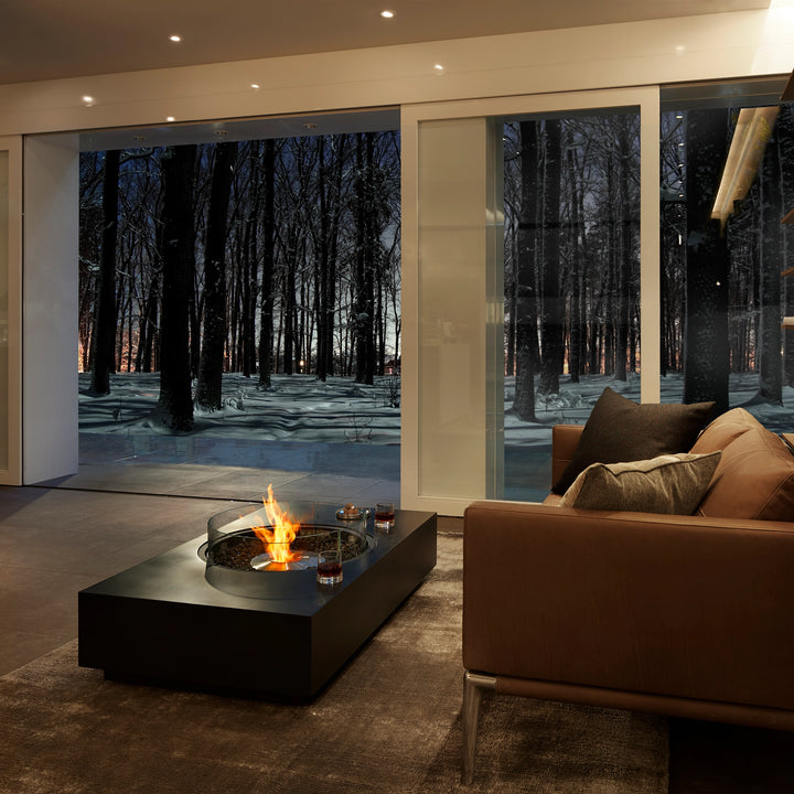 A stunning EcoSmart Fire Martini 50 gas fire table, positioned in a warm indoor setting overlooking a snowy forest. This modern fire pit with a glass wind guard provides a cozy ambiance for stylish winter interiors.