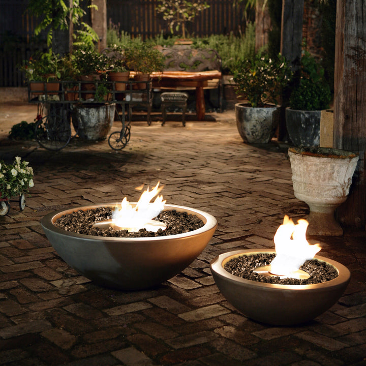 The EcoSmart Fire Mix 600 Natural Fire Pits are displayed at nighttime, casting a mesmerizing glow on a rustic brick patio. Two different-sized fire bowls with bioethanol flames provide an elegant and modern fire pit ambiance. The soft light enhances the outdoor setting, making it ideal for backyard gatherings and stylish outdoor firepit decor.