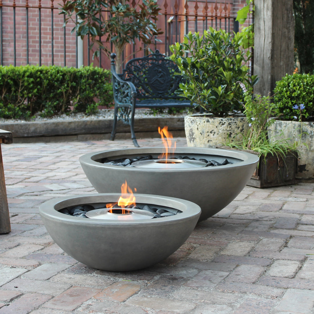 A pair of EcoSmart Fire Mix 600 Natural Fire Pits are showcased in an outdoor stone-paved patio, creating a warm and inviting atmosphere. The large and small concrete fire pits feature sleek round designs, filled with black stones and powered by bioethanol flames. These modern outdoor fire pits blend seamlessly into any outdoor fire bowl setup, providing warmth and ambiance.