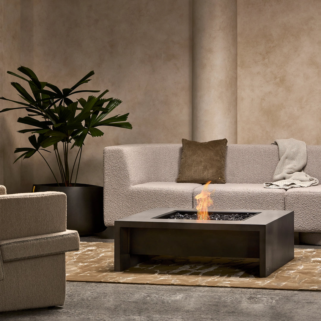 A luxury modern fire pit setup featuring the EcoSmart Fire Mojito 40 in a natural concrete finish. Positioned in a cozy indoor-outdoor living space, this square outdoor fire pit is complemented by a contemporary sofa set, a neutral-toned area rug, and an elegant black planter with lush greenery. The fire pit table enhances the ambiance with a warm, inviting flame, making it a perfect centerpiece for luxury outdoor patios, backyard fire pit setups, and contemporary lounge spaces.