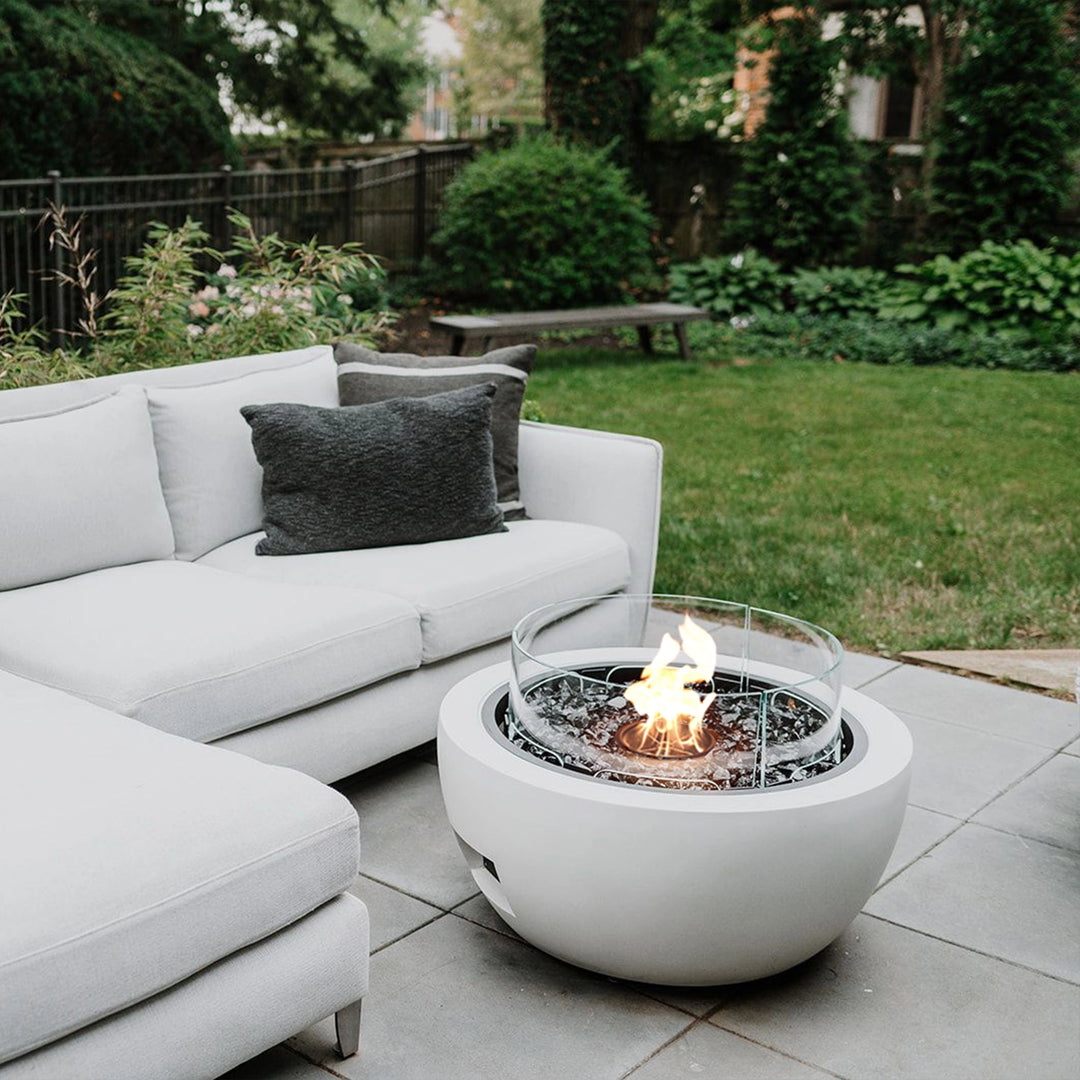 A contemporary EcoSmart Fire Pod 30 round fire bowl in a crisp white finish, surrounded by a glass wind guard, glowing with a warm flame on a modern patio seating area. Perfect for enhancing an outdoor living space, this round gas fire pit adds a luxury backyard fire feature.