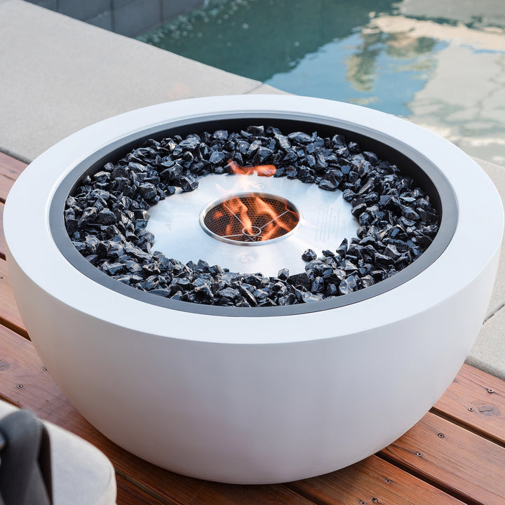 A modern EcoSmart Fire Pod 30 round fire pit in a sleek white finish, filled with black fire rocks and a stainless steel burner, elegantly placed on a wooden deck beside a swimming pool. This contemporary outdoor fire bowl provides a luxury fire pit experience, perfect for a backyard or patio setting.