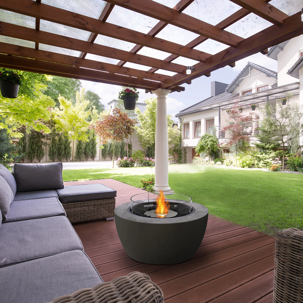 A beautifully arranged outdoor fire pit setting featuring the EcoSmart Fire Pit Pod 40 in a shaded pergola-covered patio. The round concrete fire bowl with a wind guard and black burner creates a warm and inviting atmosphere, blending seamlessly with the natural wood decking and lush green surroundings. This modern fire pit enhances the comfort of the cozy outdoor seating area, making it a perfect choice for relaxation and entertaining.