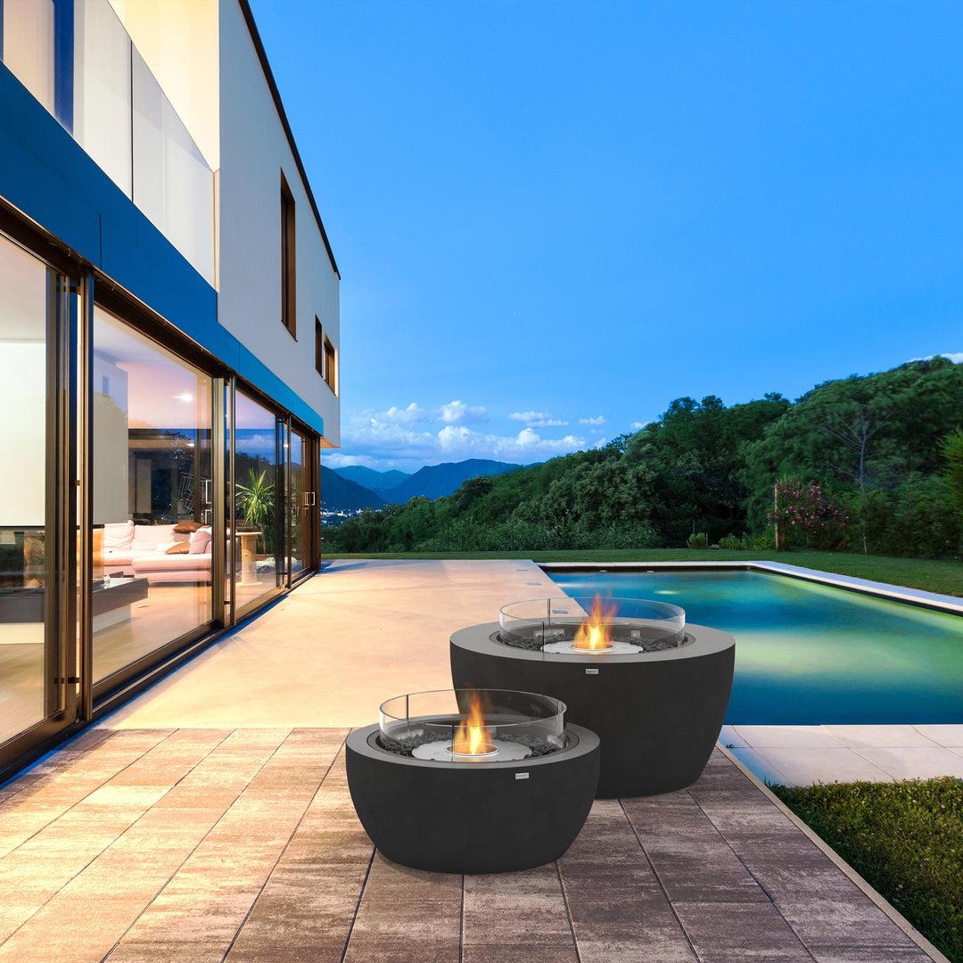  A stunning modern outdoor setting featuring two EcoSmart Fire Pit Pod 40 fire bowls placed poolside. These sleek, concrete fire pits with glass wind guards provide a mesmerizing flame display, creating a luxurious ambiance for outdoor entertaining. The dark gray finish complements the contemporary patio design, making it a stylish focal point for any backyard fire pit setup. Perfect for enhancing outdoor living spaces with a modern outdoor fire pit aesthetic.