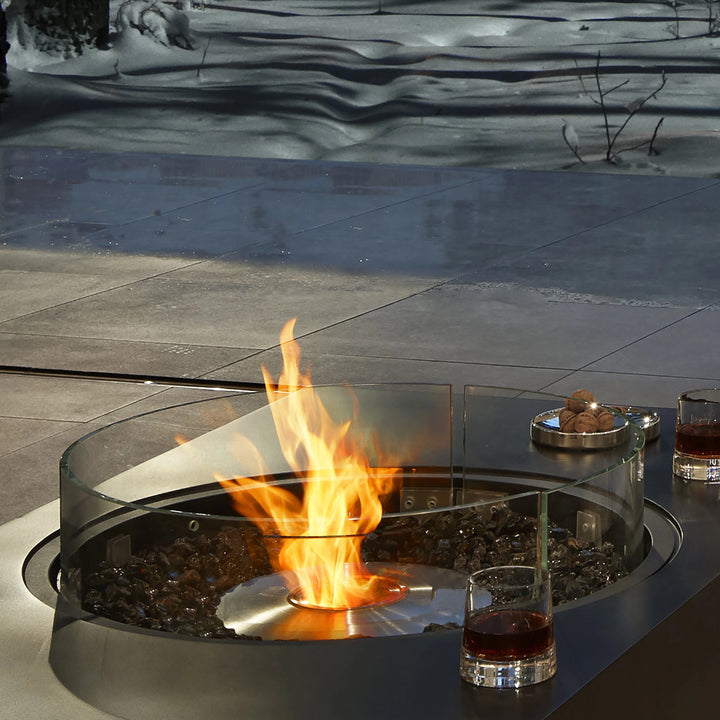 A close-up of the EcoSmart Fire R530 Fire Screen protecting the flames of a modern outdoor fire pit on a snowy patio. The sleek tempered glass wind guard enhances safety while maintaining an unobstructed view of the fire, making it an ideal outdoor fire pit accessory for cold-weather use