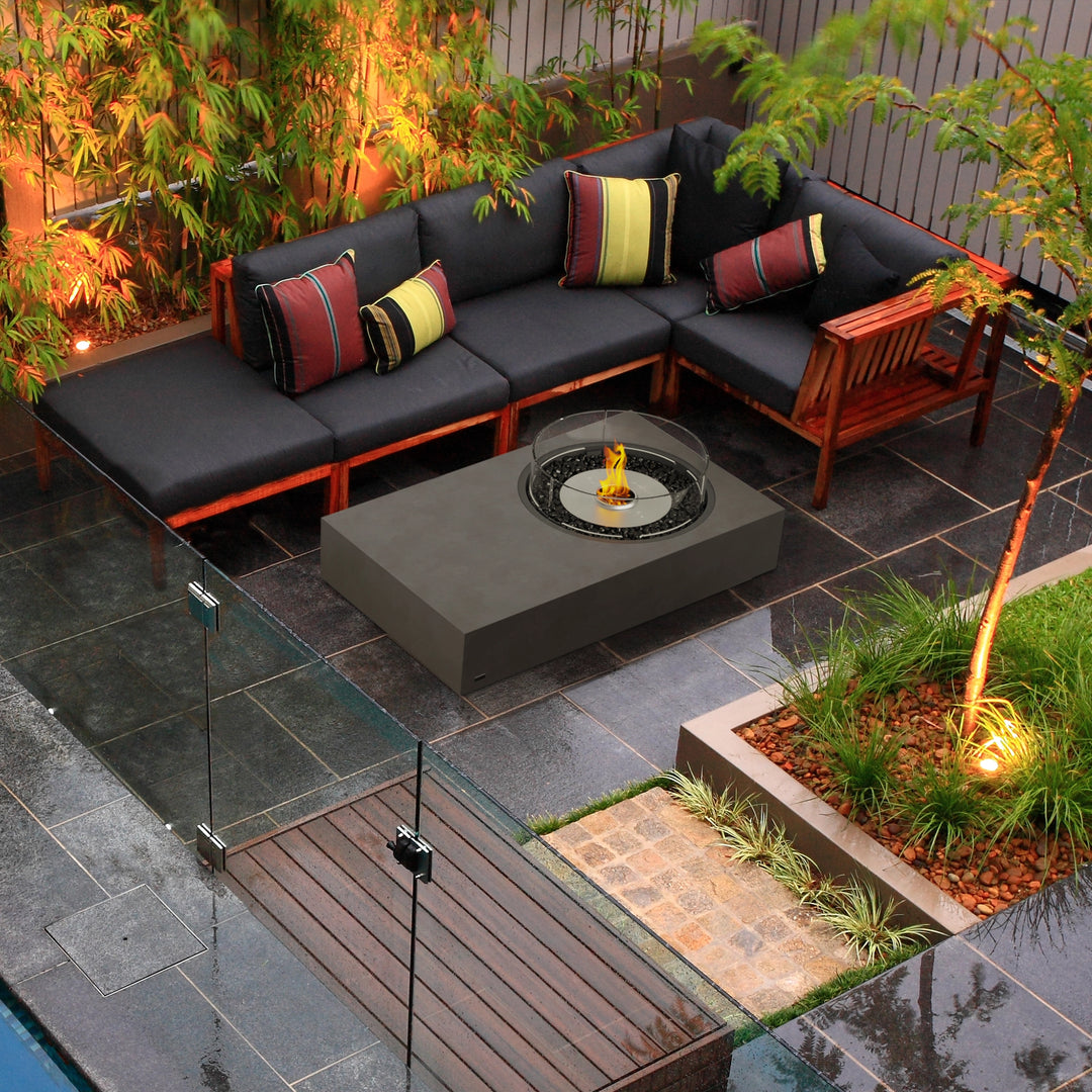 A contemporary backyard setting featuring the EcoSmart Fire Tequila 50 fire pit in a deep gray tone, surrounded by a dark cushioned L-shaped sectional with vibrant striped pillows. Positioned on a polished stone patio with lush greenery and warm lighting, the rectangular gas fire pit offers a luxurious feel. The protective glass wind guard ensures safety while maintaining an elegant look. Ideal for modern fire pits outdoor, outdoor fire tables propane, and gas fire pits outdoor.