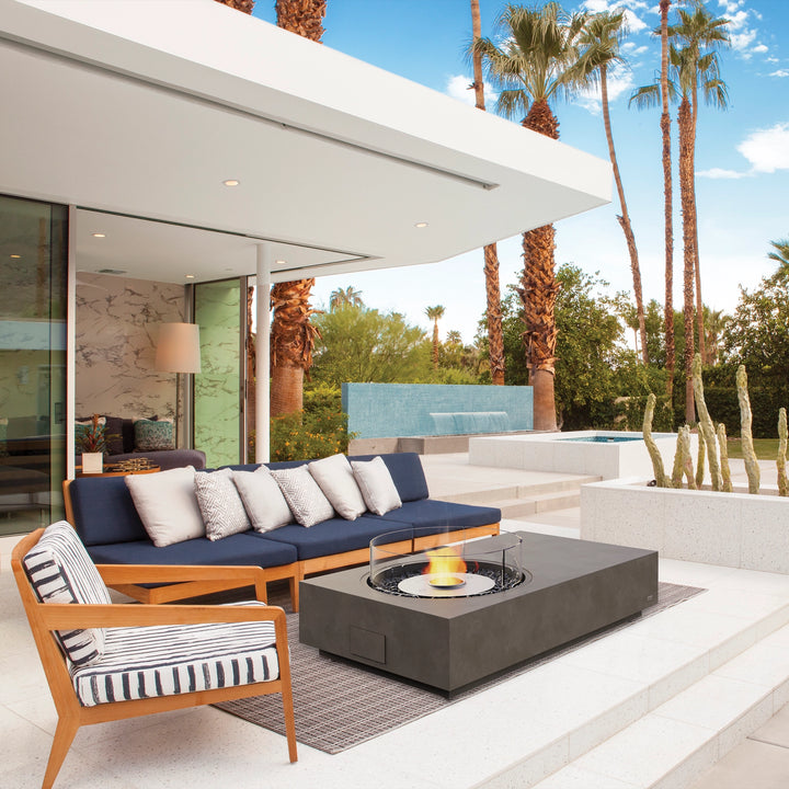 A contemporary backyard setting featuring the EcoSmart Fire Tequila 50 fire pit in a deep gray tone, surrounded by a dark cushioned L-shaped sectional with vibrant striped pillows. Positioned on a polished stone patio with lush greenery and warm lighting, the rectangular gas fire pit offers a luxurious feel. The protective glass wind guard ensures safety while maintaining an elegant look. Ideal for modern fire pits outdoor, outdoor fire tables propane, and gas fire pits outdoor.