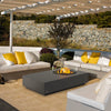 A stylish EcoSmart Fire Tequila 50 fire pit in dark gray under a pergola, surrounded by plush white and yellow-cushioned sectional seating. The large outdoor gas fire pit features a modern concrete base with a built-in burner and glass wind guard, creating an inviting focal point for social gatherings. Set against a stone wall with lush landscaping, this outdoor fire bowl enhances any contemporary patio design. Keywords: gas fire pit table outdoor, fire pit for outdoors, fire tables for patio.