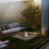 EcoSmart Fire Vertigo 50 lifestyle setting featuring a sleek modern outdoor fire pit with a natural concrete finish, surrounded by a cozy seating arrangement with a wooden sofa and armchair. The fire pit’s clean lines, glass wind guard, and glowing flames create a stylish and functional focal point for any contemporary patio or backyard space.