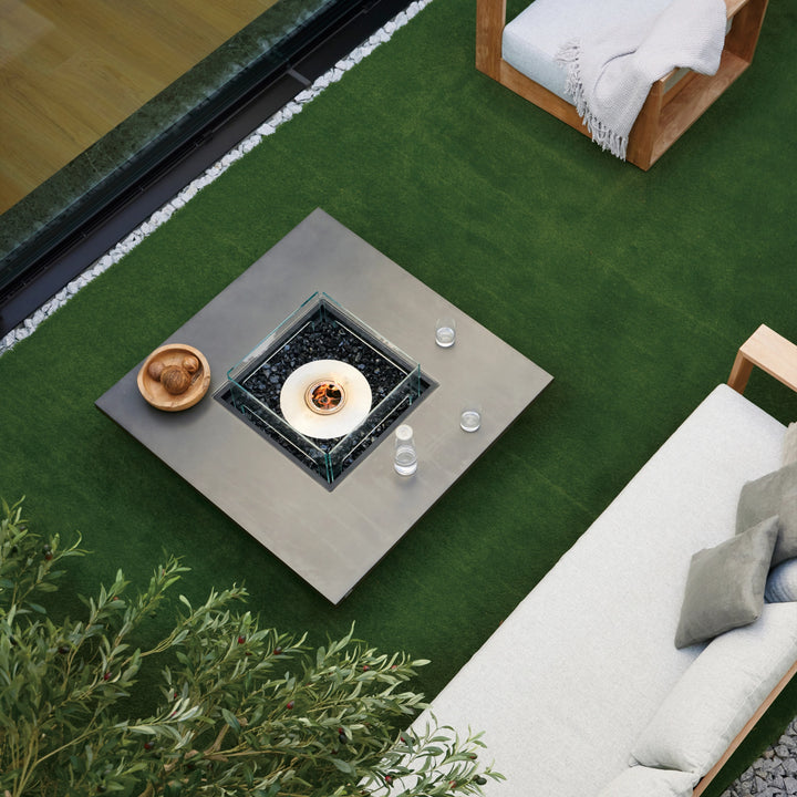 Top view of the EcoSmart Fire Vertigo 50 outdoor fire table, showcasing its spacious square design with a glass wind guard and black fire media. Positioned on artificial grass, this contemporary fire pit is surrounded by a cozy seating arrangement with a neutral-toned sofa and wooden armchairs, creating an inviting atmosphere for entertaining or relaxation