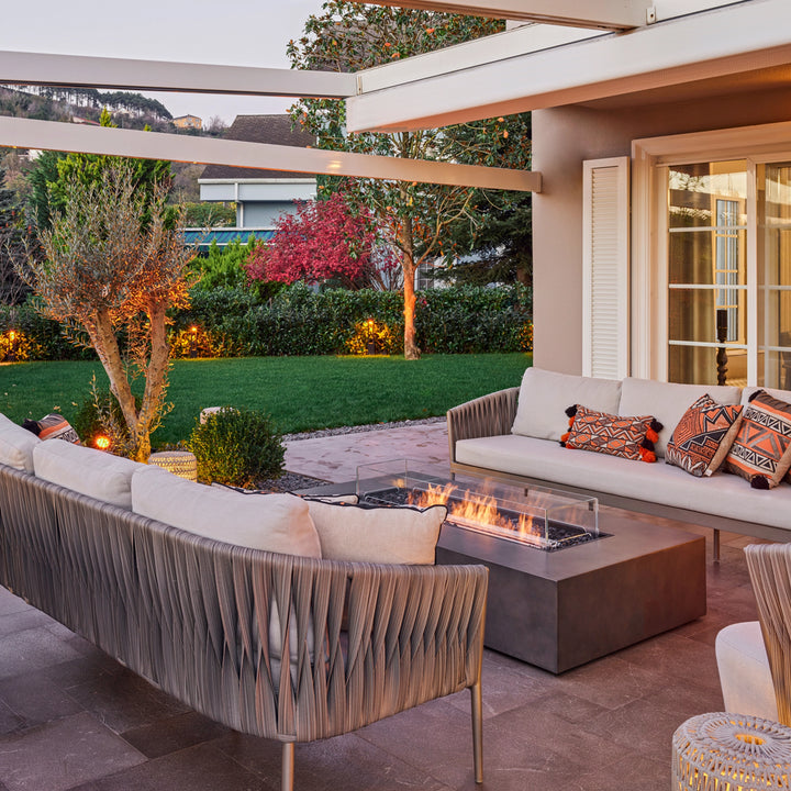 A beautifully designed garden patio featuring the EcoSmart Fire Wharf 50 in a natural concrete finish. Surrounded by plush outdoor sofas and decorative pillows, this rectangular fire table enhances the cozy ambiance, making it a perfect backyard fire pit for entertaining and relaxation.
