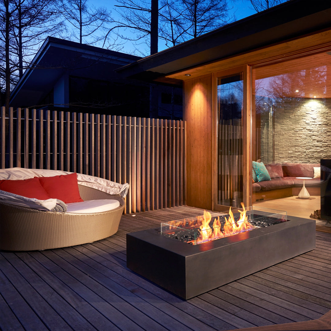 A modern outdoor deck featuring the EcoSmart Fire Wharf 50 in a graphite finish, elegantly complementing a luxurious seating area. The rectangular fire pit with a glass wind guard and black burner provides warmth and ambiance, making it a perfect outdoor fire table for modern patio settings.
