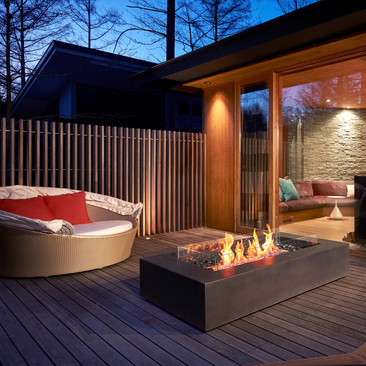 A modern outdoor deck featuring the EcoSmart Fire Wharf 50 in a graphite finish, elegantly complementing a luxurious seating area. The rectangular fire pit with a glass wind guard and black burner provides warmth and ambiance, making it a perfect outdoor fire table for modern patio settings.
