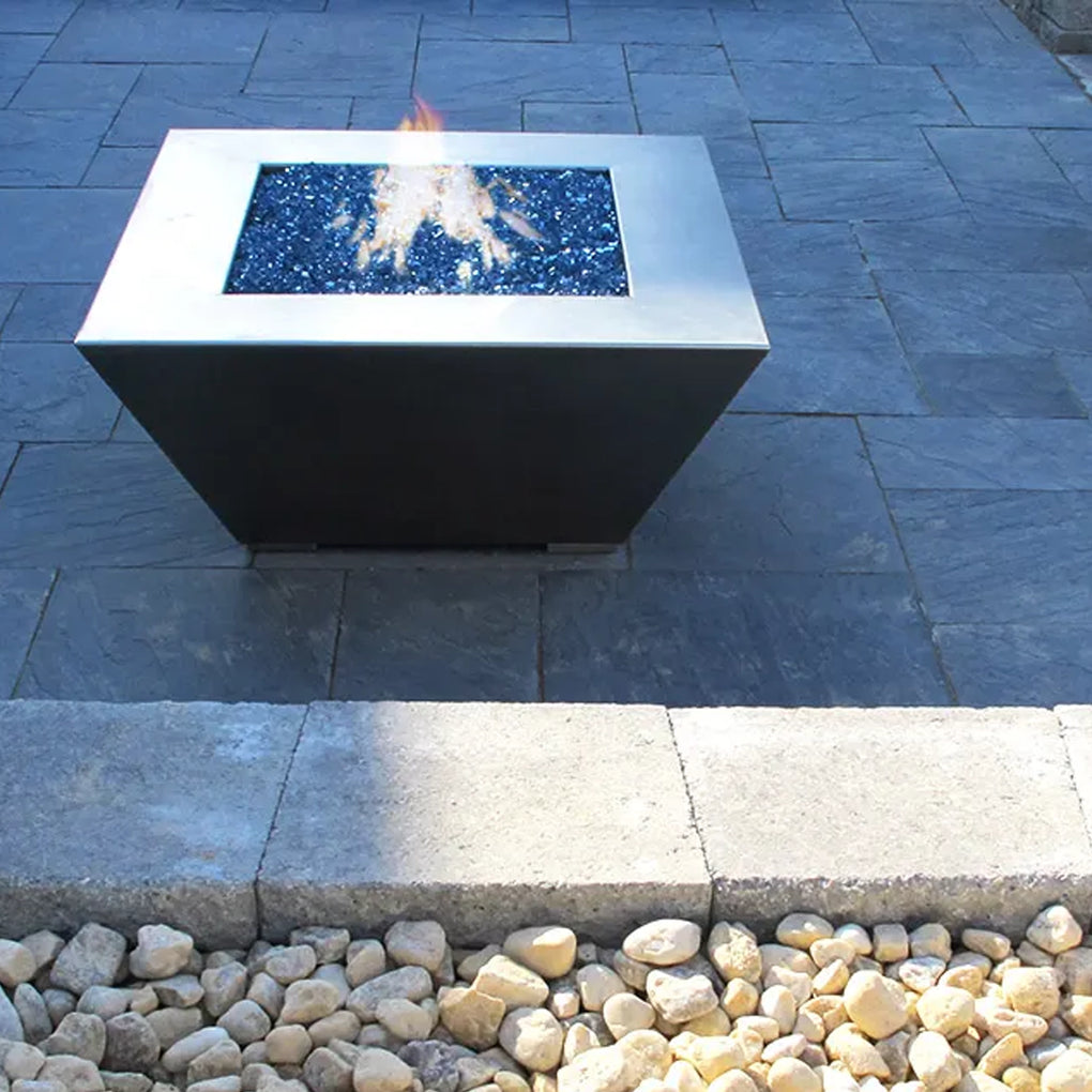 Square modern fire pit with blue fire glass on a slate patio, bordered by stone pavers and decorative gravel, creating a minimalist outdoor setting