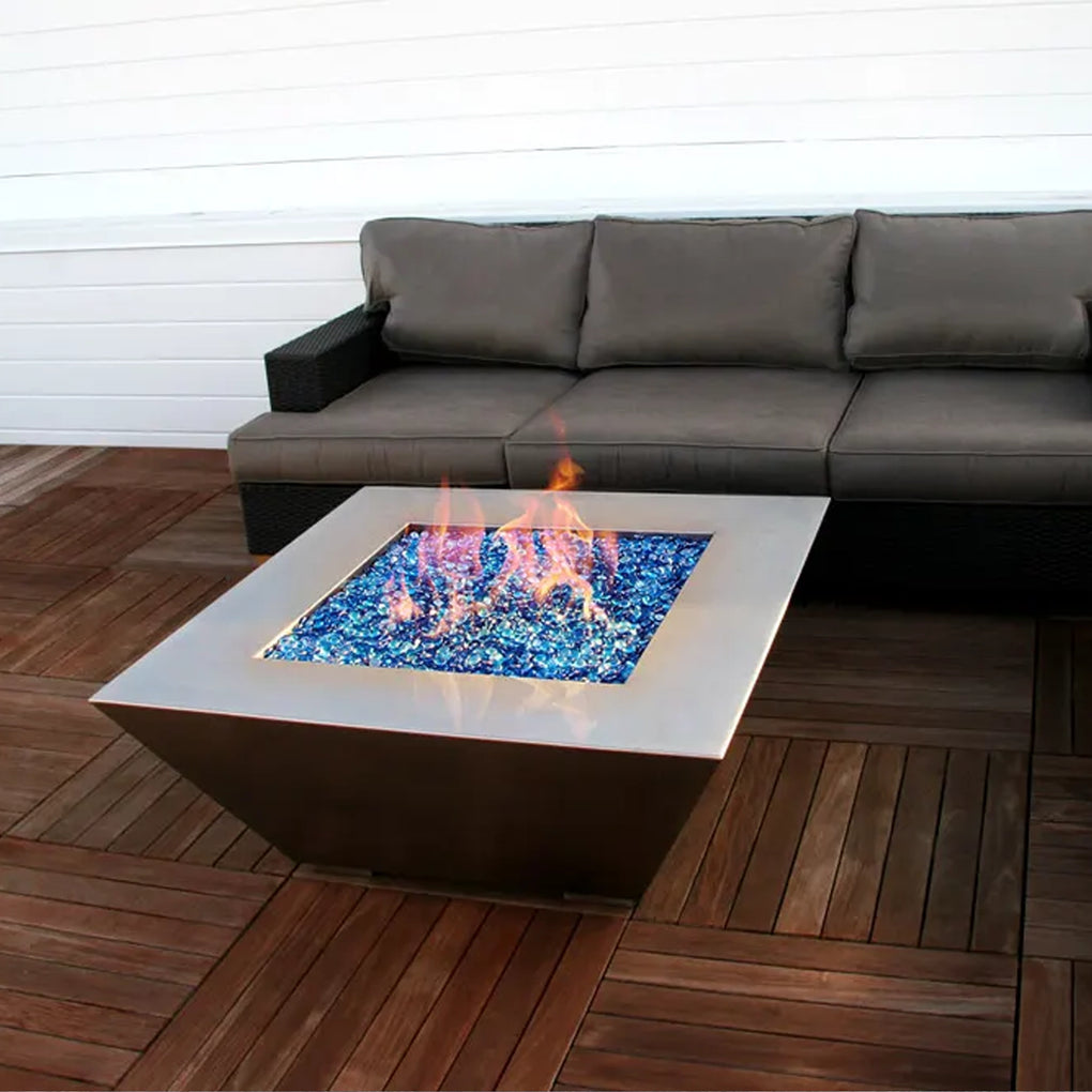 Square contemporary fire pit with blue fire glass on a wooden deck, accompanied by a dark outdoor sofa for a modern and cozy outdoor seating area.