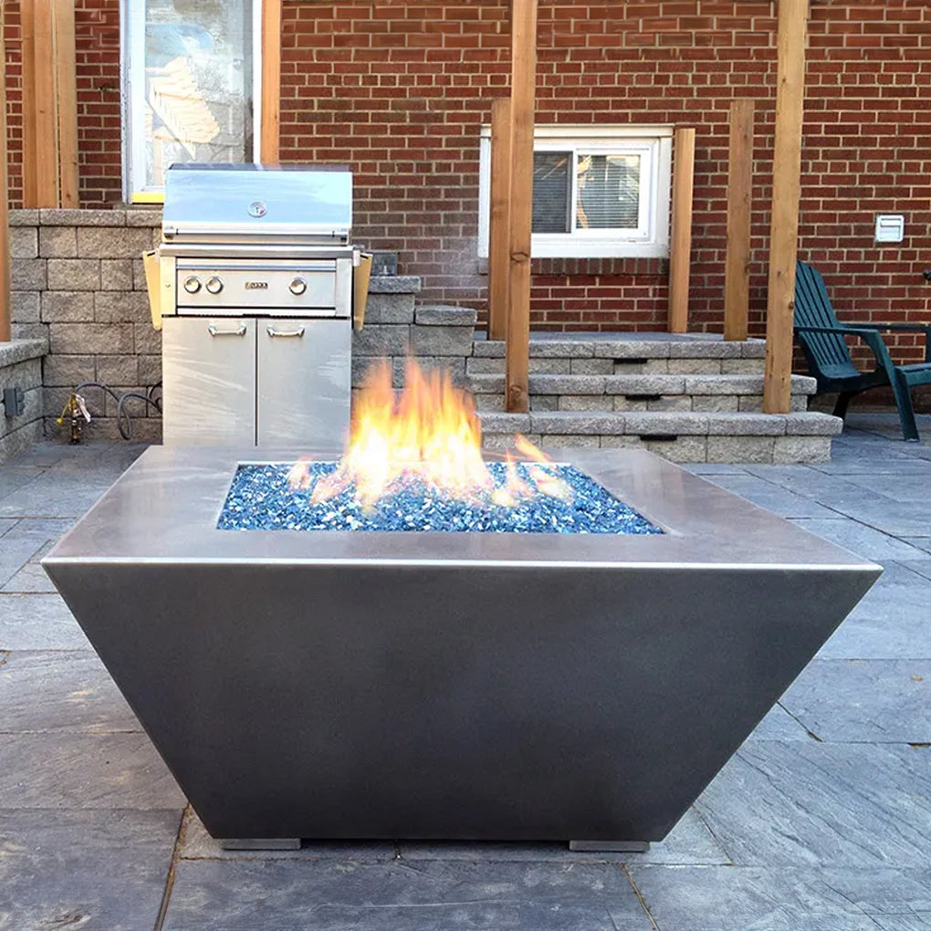 Modern square fire pit with blue fire glass on a stone patio, accompanied by a stainless steel outdoor grill, brick walls, and steps leading to a higher level in a cozy outdoor kitchen setting