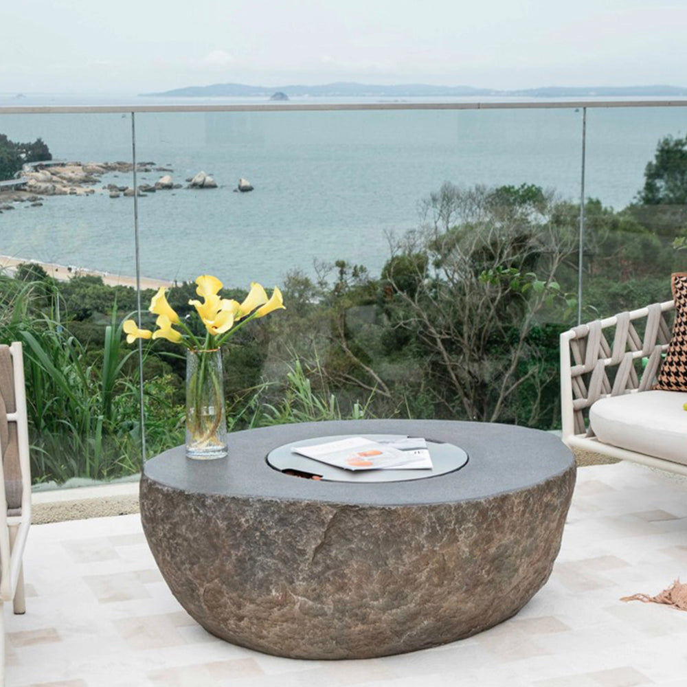 Stylish Elementi Boulder Bowl Burner Cover placed on a stone fire pit in a luxurious outdoor setting overlooking the ocean. This stainless steel cover not only provides protection but also serves as a flat surface, allowing the fire pit to double as a table when not in use. Perfect for enhancing outdoor patio aesthetics.