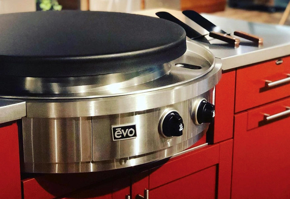 Evo stainless steel flat-top grill with black cooking surface, installed in an outdoor kitchen with red cabinetry and grilling utensils on the countertop.