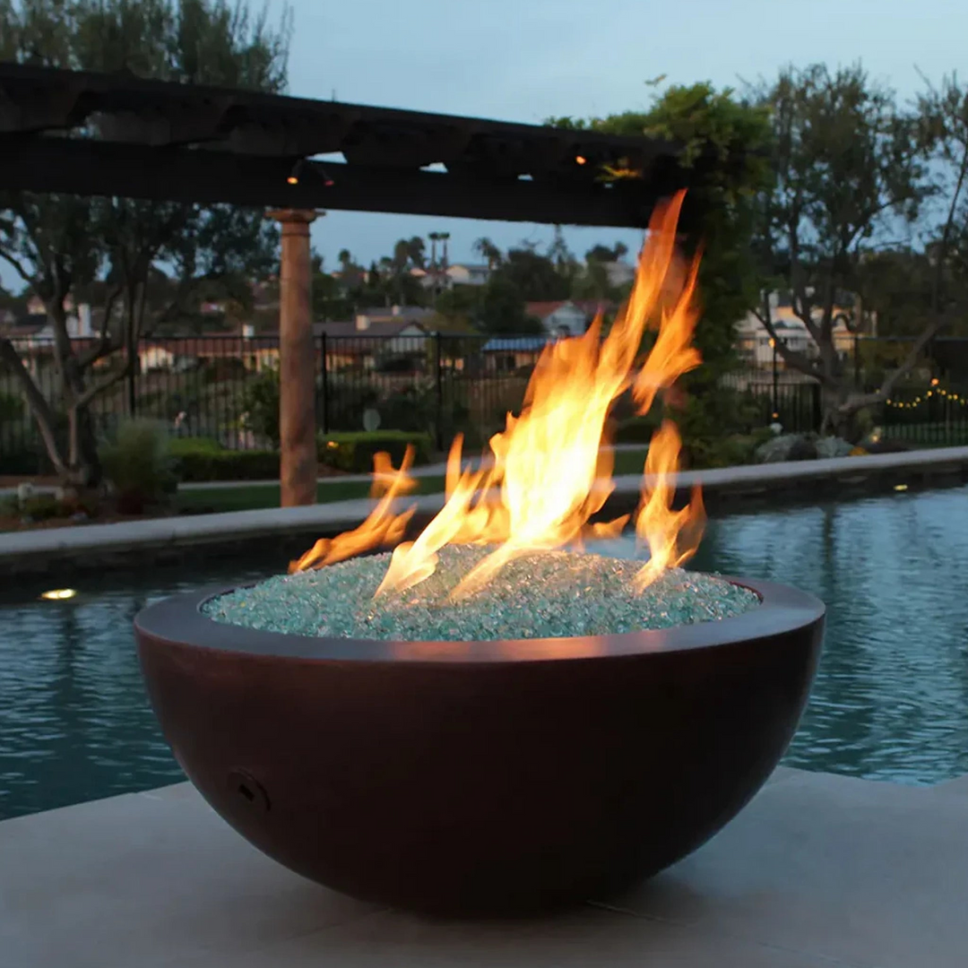 Outdoor round copper fire pit filled with reflective fire glass, placed by a swimming pool with bright flames, set against a backdrop of a landscaped garden and pergola at dusk