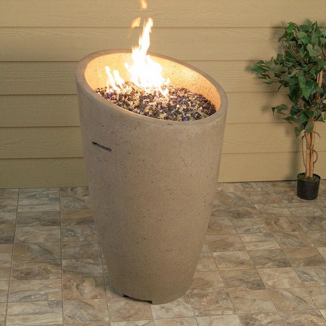 Tall cone-shaped beige outdoor fire pit with a slanted opening, filled with lava rocks and a burning flame, placed on a tiled patio next to a beige siding wall and a small potted plant