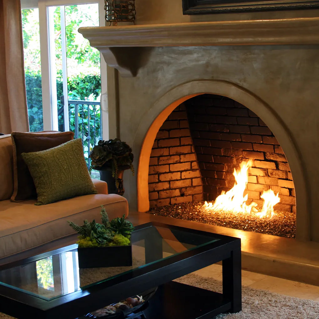 Cozy living room featuring an arched brick fireplace with glowing flames, adorned with glass fire pit media, a beige sofa with green pillows, and a glass coffee table with a succulent plant