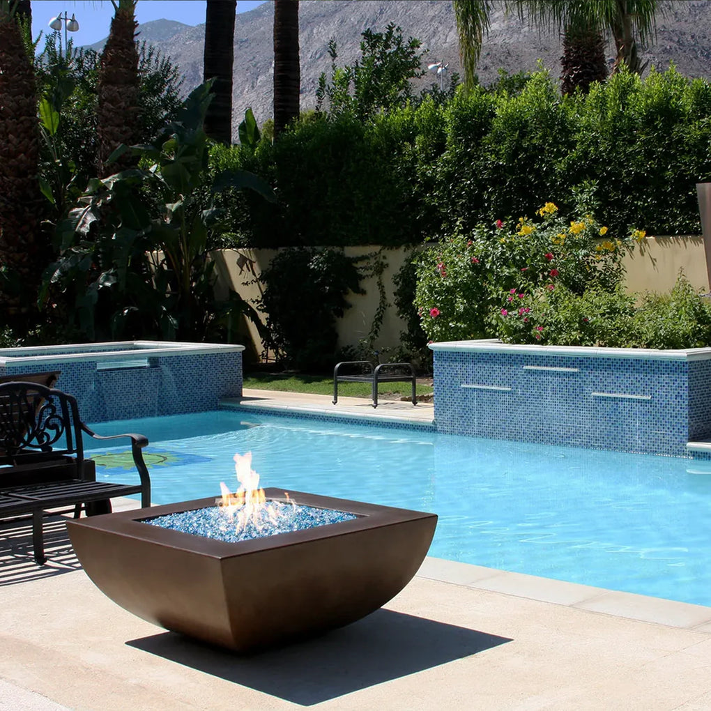 Square bronze fire bowl filled with blue fire glass, emitting flames next to a luxurious swimming pool in a tropical outdoor setting