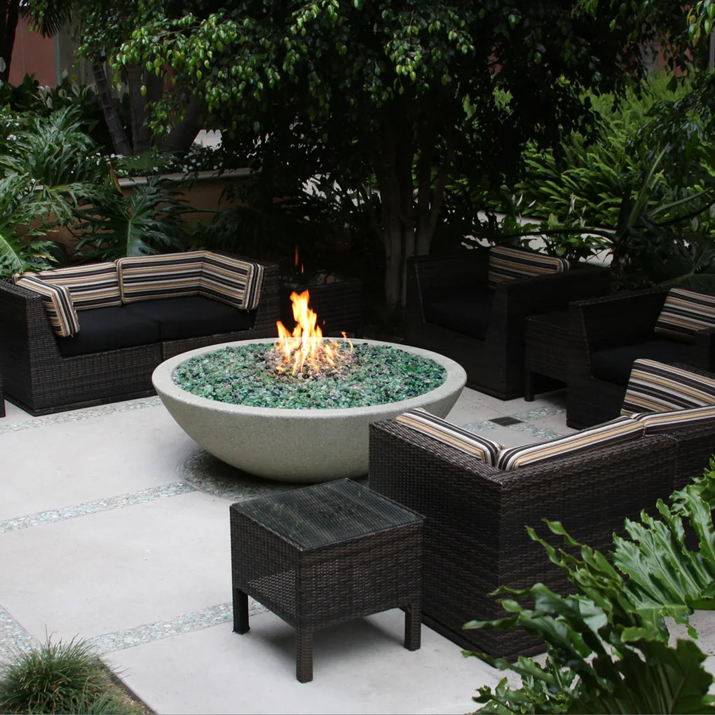 Round concrete fire bowl filled with green fire glass, emitting flames in a cozy outdoor patio setting with rattan furniture and lush greenery