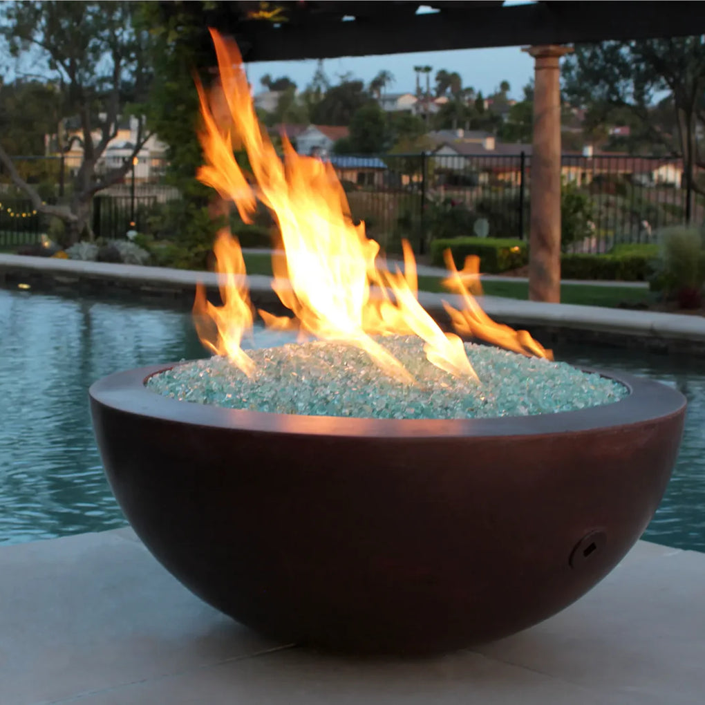 Round copper fire bowl filled with aqua fire glass, producing a warm flame next to a luxurious poolside setting at dusk