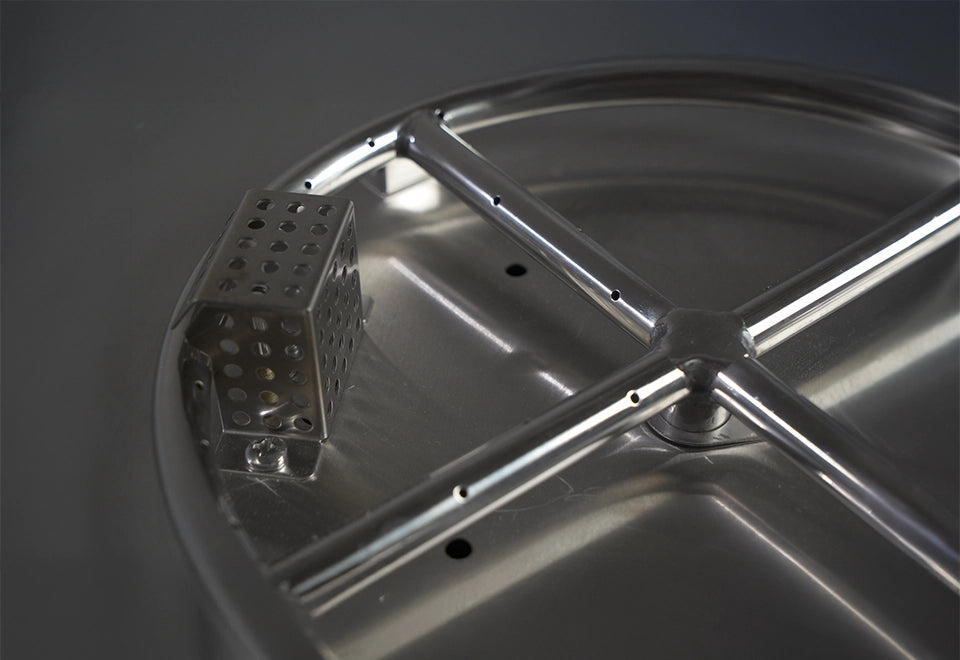 Close-up of a stainless steel fire pit burner with precision-drilled holes for even flame distribution and a protective igniter cage.